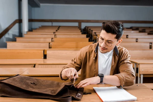 Focused male student in glasses sitting at desk and looking for pen in leather bag during lesson in classroom — Stock Photo