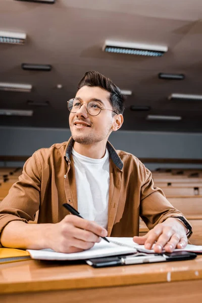 Smiling male student in glasses sitting at desk and writing in notebook during lesson in classroom — Stock Photo