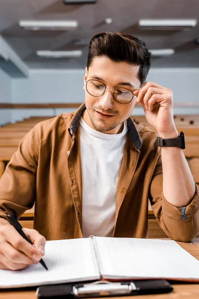 Concentrated male student in glasses sitting at desk and writing in notebook during lesson in classroom — Stock Photo