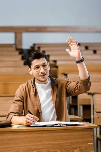 Bello sorridente studente maschio in occhiali seduto alla scrivania e alzando la mano durante la lezione in classe — Foto stock