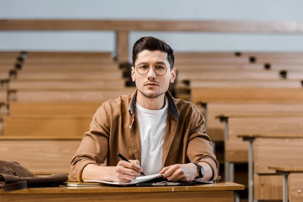 Focused male student in glasses sitting at desk, looking at camera while writing in notebook during lesson in classroom — Stock Photo