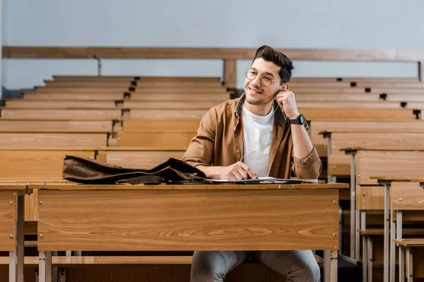 Усміхнений студент чоловічої статі в окулярах сидить за столом і дивиться в очі в класі — стокове фото