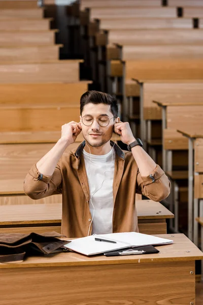 Male student in glasses sitting at desk and putting on earphones in classroom — Stock Photo