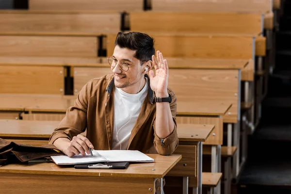 Lächelnder männlicher Student mit Brille sitzt am Schreibtisch und hebt während des Unterrichts im Klassenzimmer die Hand — Stockfoto