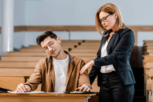 Hochschullehrerin schaut männlichen Studenten mit Brille bei schriftlicher Prüfung im Hörsaal an — Stockfoto