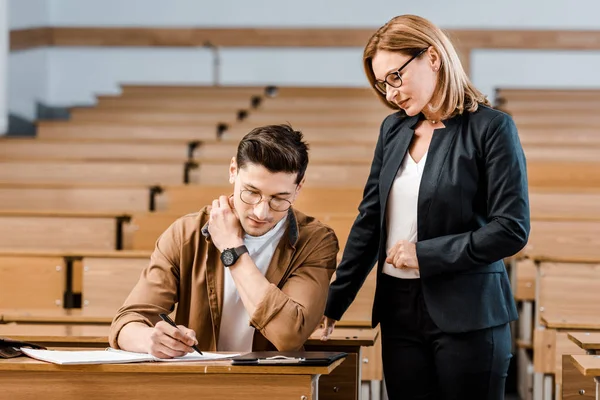 Female university teacher looking at male student writing exam in classroom — Stock Photo