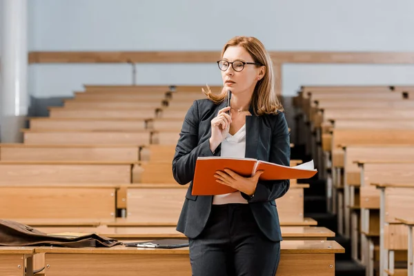 Pensativa profesora universitaria sosteniendo diario y mirando hacia otro lado en el aula - foto de stock