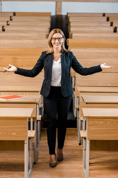 Souriante professeure d'université avec les mains tendues regardant la caméra dans la salle de classe — Photo de stock