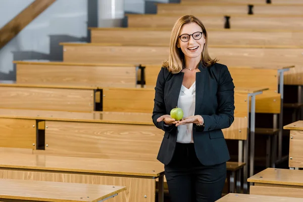 Profesora sonriente en gafas y ropa formal sosteniendo manzana y mirando a la cámara en el aula - foto de stock