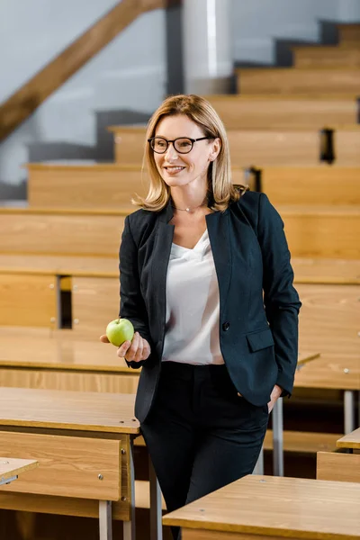 Smiling female teacher in formal wear holding apple in classroom — Stock Photo
