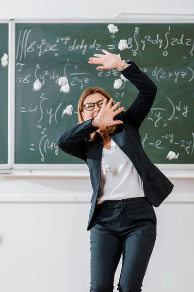 Crumpled paper balls flying at frightened female teacher in classroom with chalkboard on background — Stock Photo