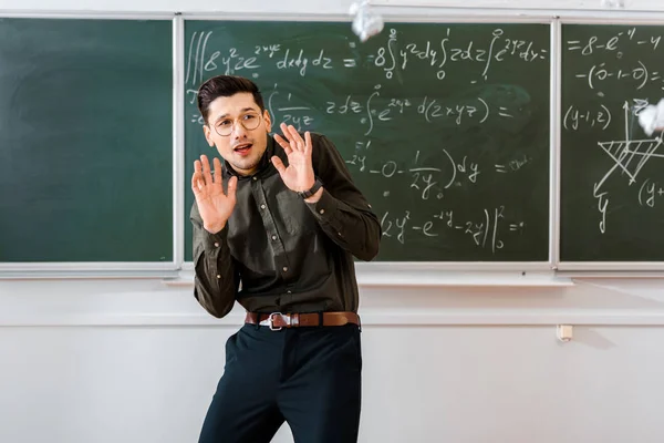 Bolas de papel amassadas voando em assustado professor masculino em sala de aula com quadro-negro no fundo — Fotografia de Stock