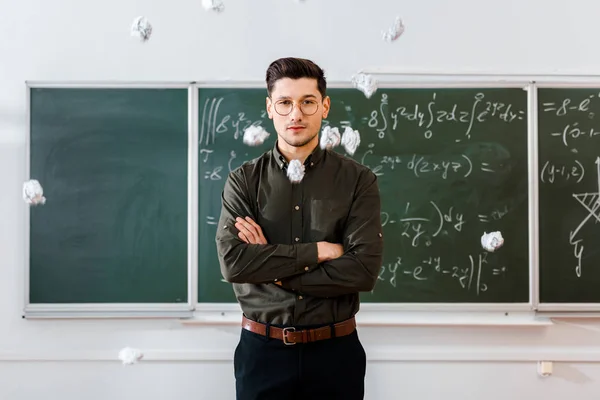 Crumpled paper balls flying at male teacher with arms crossed in classroom with chalkboard on background — Stock Photo