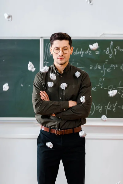 Crumpled paper balls flying at male teacher with arms crossed in classroom with chalkboard on background — Stock Photo