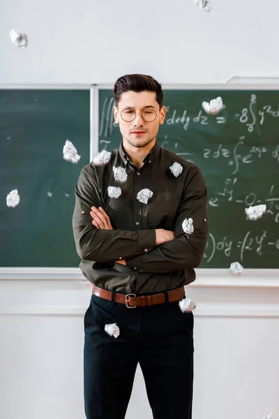 Bolas de papel arrugado volando a profesor masculino con los brazos cruzados en el aula con pizarra en el fondo - foto de stock