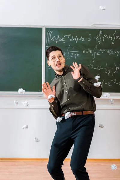 Crumpled paper balls flying at frightened male teacher in classroom with chalkboard on background — Stock Photo