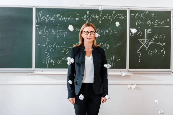 Crumpled paper balls flying at female teacher in classroom with chalkboard on background — Stock Photo