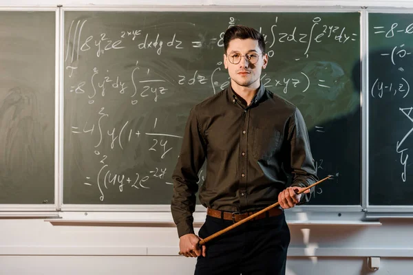 Focused male teacher in formal wear looking at camera and holding wooden pointer in front of chalkboard with equations — Stock Photo