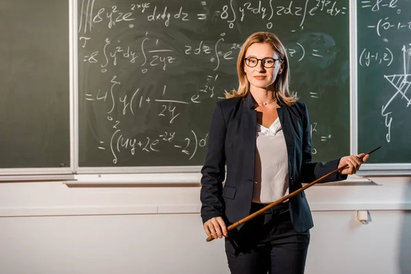 Professora atraente com ponteiro de madeira explicando equações matemáticas em sala de aula — Fotografia de Stock