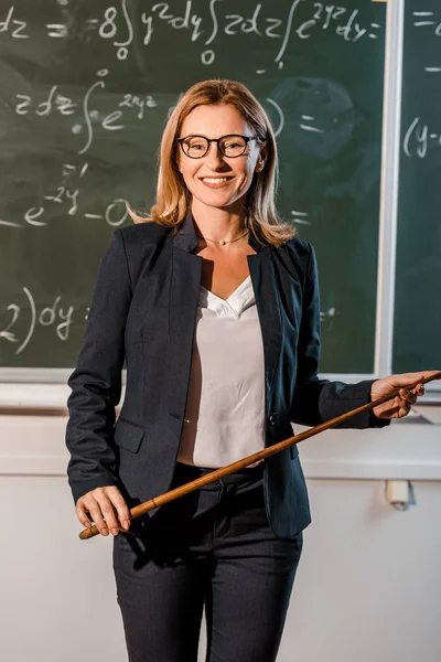 Insegnante donna in abito formale con puntatore in legno che spiega equazioni matematiche in classe — Foto stock