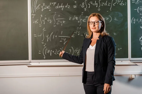 Bella insegnante femminile con puntatore in legno che spiega equazioni matematiche in classe — Foto stock