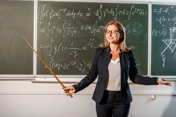 Улыбающаяся учительница с деревянным указателем, объясняющим математические уравнения в классе — стоковое фото