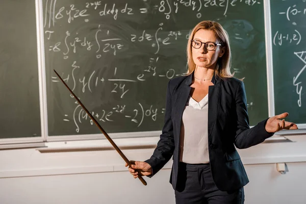 Enseignante en tenue formelle avec pointeur en bois expliquant les équations mathématiques en classe — Photo de stock