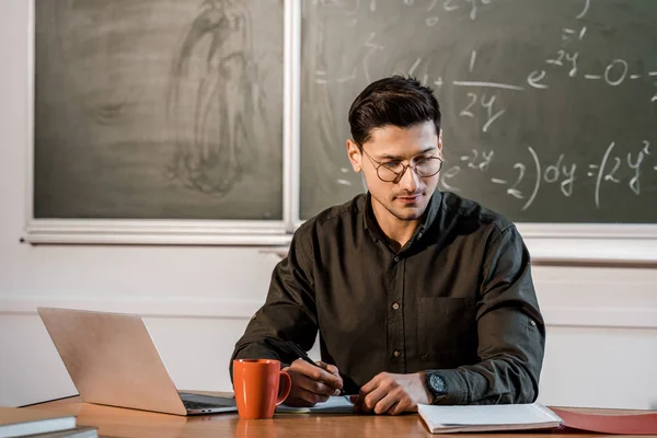 Pensive male teacher in glasses sitting at computer desk with notebooks and coffee in classroom — Stock Photo