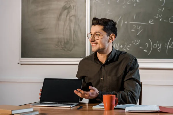Profesor masculino sonriente sentado en el escritorio y mostrando portátil con pantalla en blanco en el aula - foto de stock