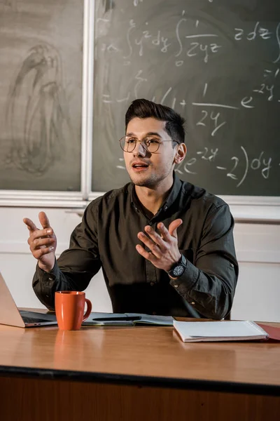 Profesor de gafas sentado en el escritorio y explicando ecuaciones en el aula de matemáticas - foto de stock