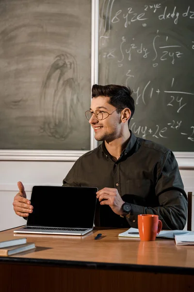 Усміхнений вчитель чоловічої статі в окулярах сидить за столом і показує ноутбук з порожнім екраном в класі — стокове фото