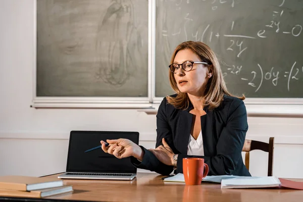 Professora sentada na mesa e apontando para laptop com tela em branco em sala de aula — Fotografia de Stock