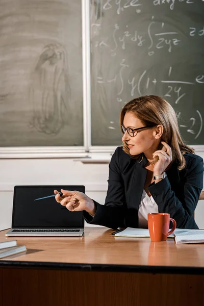 Profesora en gafas sentada en el escritorio y apuntando a la computadora portátil con pantalla en blanco en el aula - foto de stock