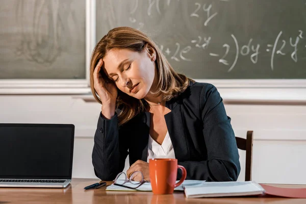 Tired female teacher with eyes closed sitting at computer desk in classroom — Stock Photo