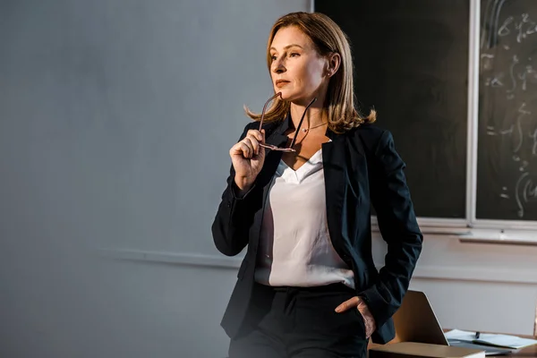 Female teacher in formal wear holding glasses and looking away in classroom — Stock Photo