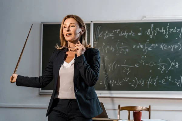 Professora com ponteiro de madeira segurando óculos e explicando equações matemáticas em sala de aula — Fotografia de Stock