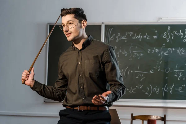 Bello maschio insegnante in abbigliamento formale tenendo puntatore di legno e spiegando equazioni in classe — Foto stock