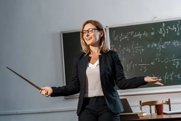 Insegnante sorridente con puntatore in legno che spiega le equazioni matematiche in classe — Foto stock