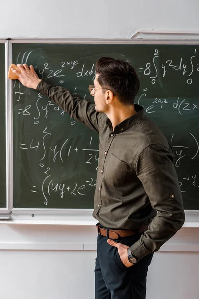 Profesor masculino limpiando ecuaciones matemáticas con esponja de pizarra en el aula - foto de stock