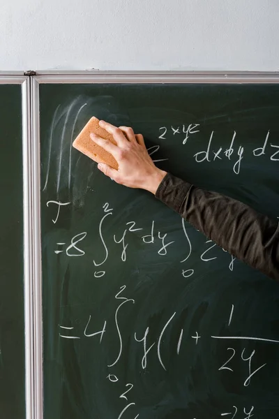 Vista recortada de estudiante masculino limpiando ecuaciones matemáticas con esponja en el aula - foto de stock