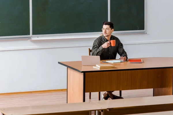 Male teacher sitting at computer desk and drinking coffee in classroom — Stock Photo