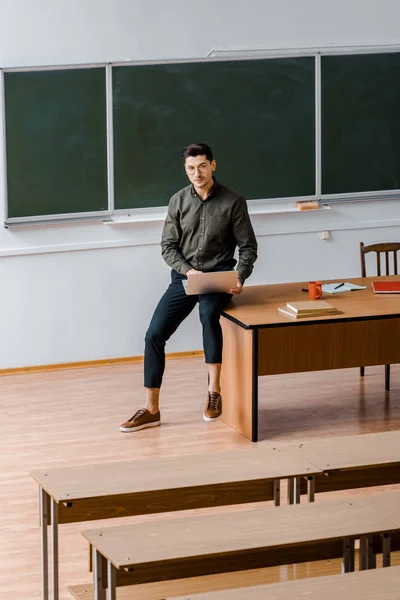 Чоловічий вчитель у формальному одязі сидить на столі і тримає ноутбук у класі — стокове фото