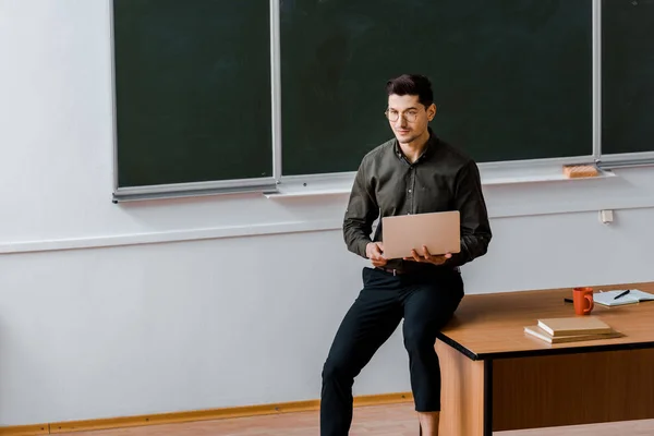 Maestro masculino en ropa formal sentado en el escritorio y sosteniendo el ordenador portátil en el aula - foto de stock