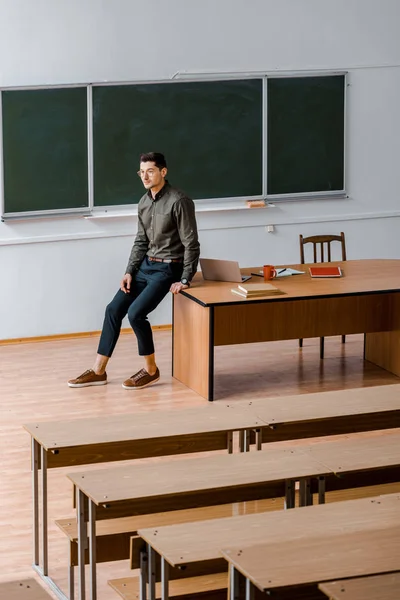 Male teacher in formal wear looking away and sitting on desk in classroom — Stock Photo