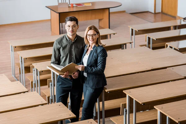 Estudiante masculino sonriente y profesora de sexo femenino sosteniendo libros universitarios y mirando a la cámara en el aula - foto de stock
