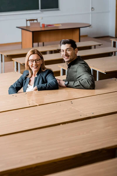 Estudante do sexo masculino e professora olhando para a câmera, sorrindo e sentado em sala de aula — Fotografia de Stock