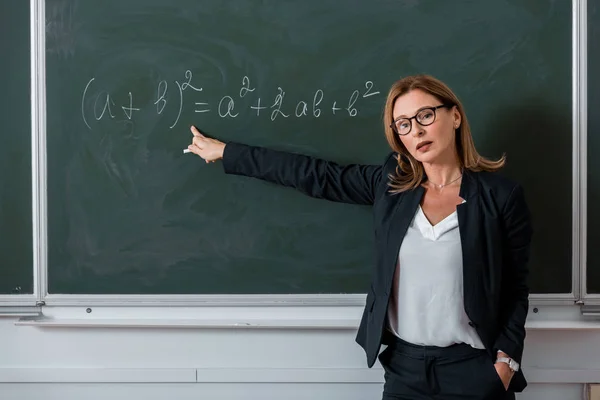 Female teacher looking at camera and pointing with finger at mathematical equation on chalkboard in class — Stock Photo