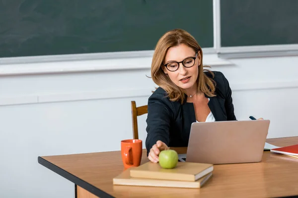 Hermosa profesora de gafas sentada en el escritorio de la computadora y buscando manzana en el aula - foto de stock