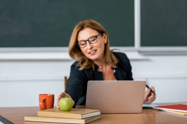 Profesora sentada en el escritorio de la computadora y buscando manzana en el aula - foto de stock