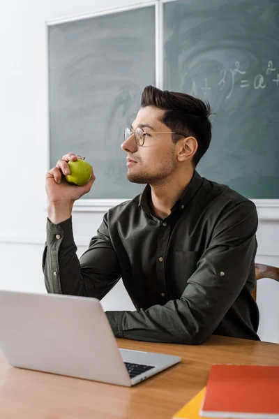 Целенаправленный учитель в очках сидит за компьютером и держит яблоко в классе — стоковое фото